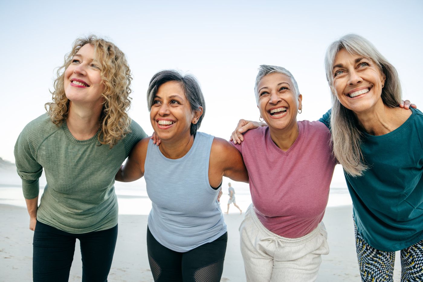 Four women in midlife walking arm-in-arm and laughing on a beach